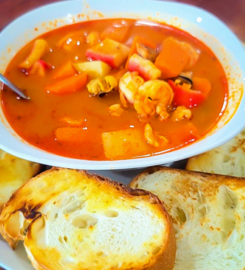 A bowl of colorful seafood soup with chunks of vegetables, served with toasted bread slices on the side.