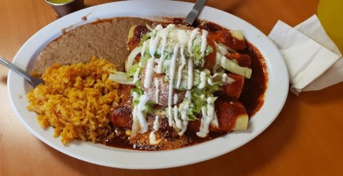 A plate of enchiladas topped with lettuce, sour cream, and cheese, served with rice and refried beans on the side.
