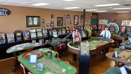 A vintage casino interior with various gaming tables, slot machines, and a roulette wheel, featuring two staff members.