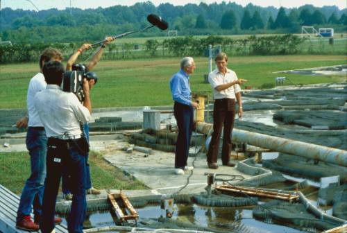 A film crew records two men discussing a large outdoor water project with pipes and equipment in the background.
