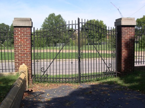 Wrought iron gate with brick pillars, opening to a grassy area and trees, under a clear blue sky.