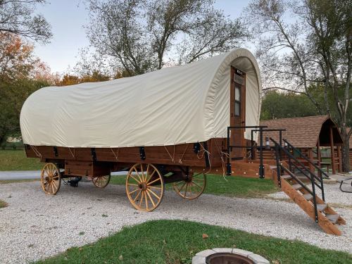 A wooden covered wagon with a white canvas top, parked on gravel near trees and a fire pit.
