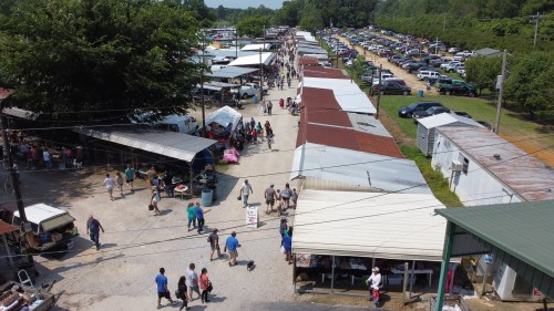Aerial view of a bustling outdoor market with vendors, shoppers, and parked cars along a tree-lined path.