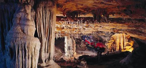 A cave interior with impressive stalactites and stalagmites, featuring a red tour vehicle and illuminated rock formations.