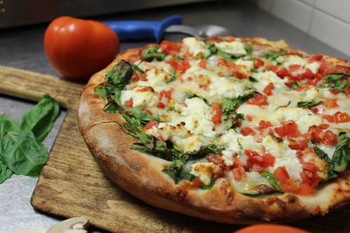 A freshly baked pizza topped with spinach, tomatoes, and cheese on a wooden cutting board, with a tomato and mushrooms nearby.