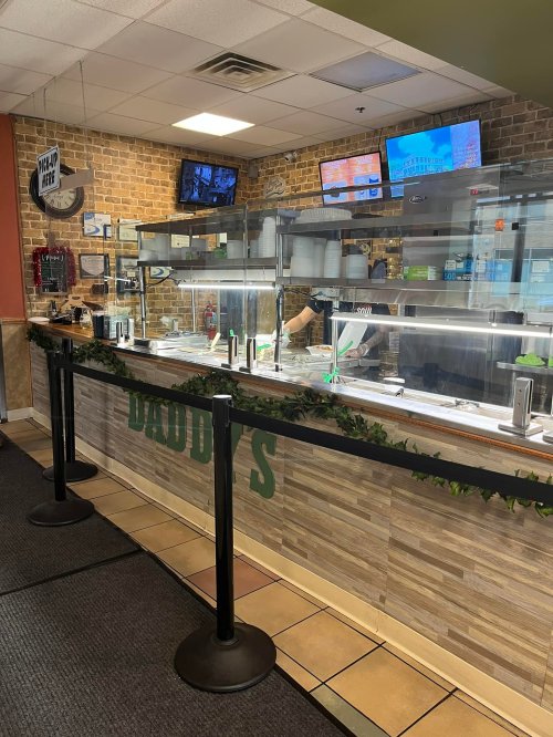 A restaurant counter with a menu display, food trays, and a staff member serving customers. Green decor along the front.
