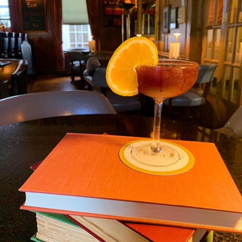A cocktail with an orange slice sits atop a stack of colorful books in a cozy bar setting.