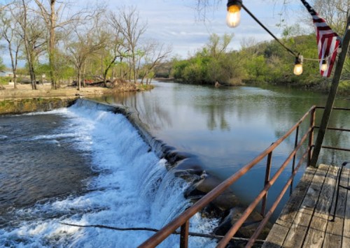 A serene river scene with a small waterfall, surrounded by trees and a wooden railing, featuring hanging lights and an American flag.