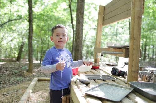 A young boy stands at a wooden outdoor table, playing with water and utensils in a forested area.