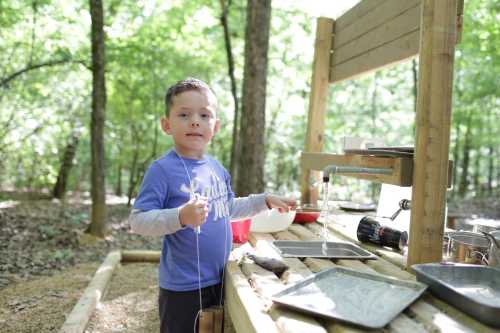 A young boy stands at a wooden outdoor table, playing with water and utensils in a forested area.