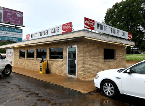 Exterior of White Trolley Cafe, a brick building with Coca-Cola signage, surrounded by a parking area and greenery.