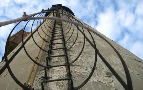 A close-up view of a rusty metal ladder spiraling up a weathered tower against a cloudy sky.