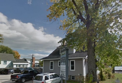 A small house with a large tree nearby, surrounded by parked cars and under a partly cloudy sky.
