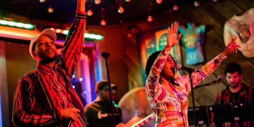 A lively music performance with two singers on stage, surrounded by colorful lights and an engaged audience.