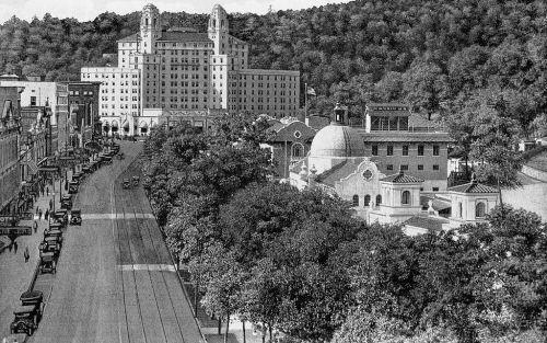 Historic black-and-white view of a hotel and surrounding buildings, with trees and early 20th-century cars on the street.