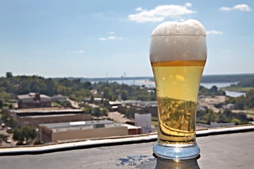 A glass of beer with a frothy head sits on a ledge, overlooking a scenic view of a town and river under a clear sky.
