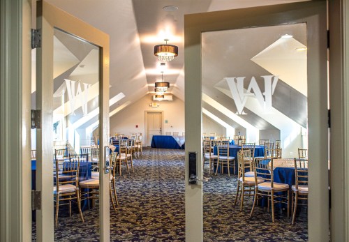 View of a banquet room through double doors, featuring round tables with blue tablecloths and elegant chandeliers.