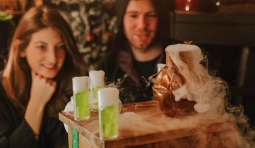 A woman and man watch as a bubbling pot emits smoke, with green drinks in front of them.