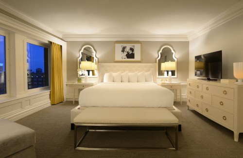 A stylish hotel room featuring a large bed, elegant decor, and large windows with city views at dusk.