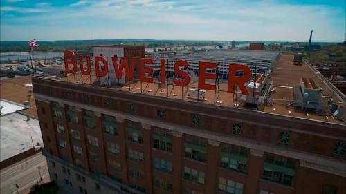 Aerial view of a large building with a prominent Budweiser sign on the roof, set against a clear blue sky.