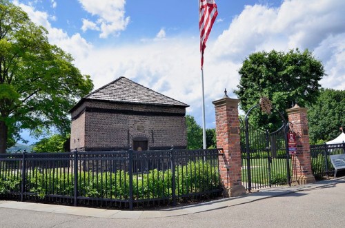 Historic brick building with a gabled roof, surrounded by a black fence and greenery, under a blue sky with clouds.