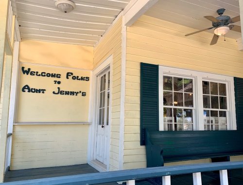 A welcoming porch with a sign reading "Welcome Folks to Aunt Jenny's" on a yellow wall and green shutters.