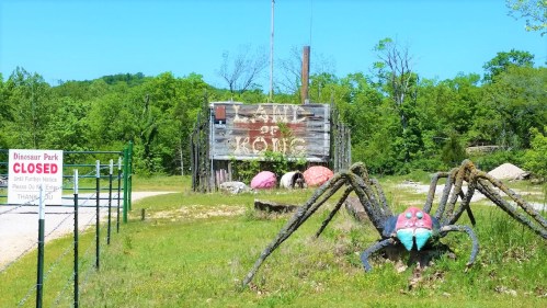 A colorful spider sculpture in front of a closed dinosaur park sign, surrounded by greenery and a rustic wooden structure.