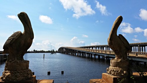 Two stone pelican statues frame a view of a bridge over a calm waterway under a blue sky with fluffy clouds.