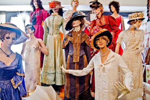 A collection of vintage mannequins dressed in elegant historical clothing and hats, displayed in a bright room.