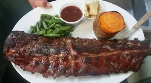 A large plate featuring ribs, green beans, a sweet potato, and a small cup of barbecue sauce.