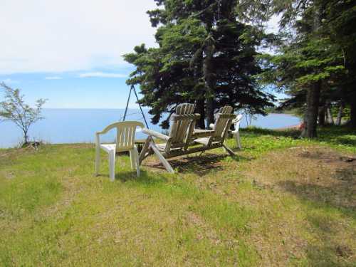 Two wooden adirondack chairs and a white plastic chair sit on a grassy area by the water, surrounded by trees.