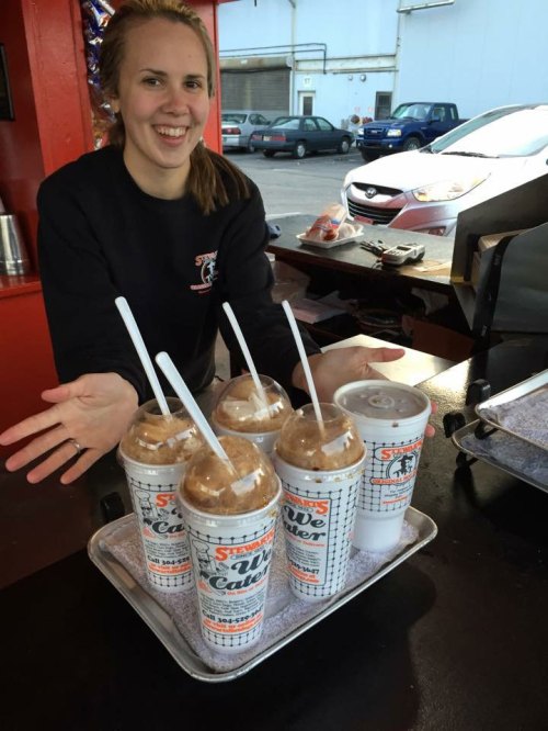 A smiling person stands behind a tray of five frosty drinks with straws, showcasing a cheerful atmosphere.