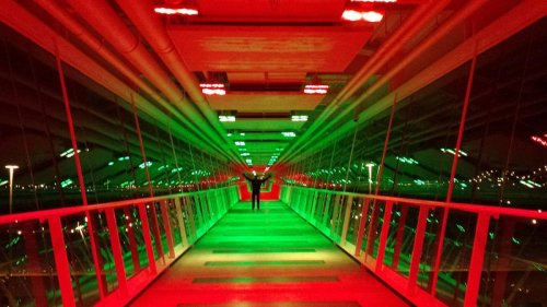 A person stands with arms outstretched in a brightly lit corridor, illuminated in red and green lights.