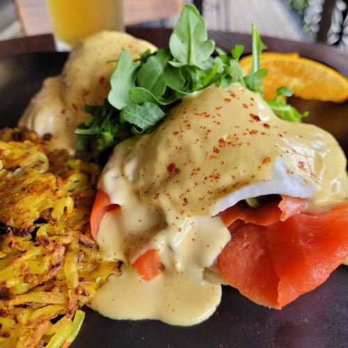 A plate featuring eggs benedict with smoked salmon, hollandaise sauce, arugula, and crispy hash browns, garnished with orange.