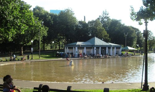 A serene park scene featuring a pond, a gazebo, and people enjoying the outdoors on a sunny day.