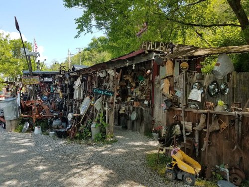 A quirky outdoor display of vintage items and tools arranged along a rustic wooden fence under green trees.