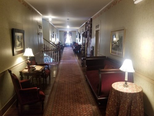 A long, elegantly decorated hallway with vintage furniture, lamps, and patterned carpet, leading to a staircase.