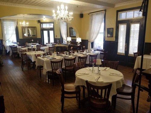 A cozy restaurant interior with wooden floors, round tables set for dining, and elegant chandeliers.