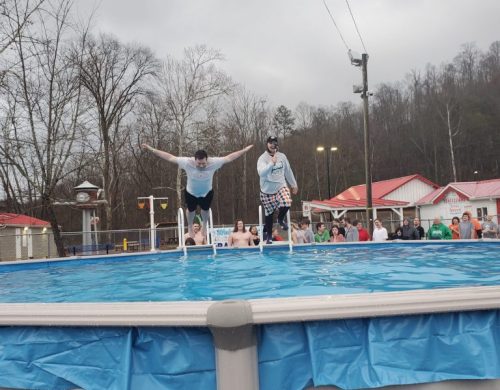 Two people jump into a pool while a crowd watches from the side on a cloudy day.
