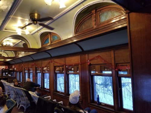 Interior of a vintage train car with wooden paneling, festive decorations, and snowy windows visible outside.