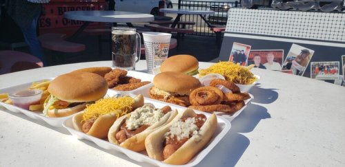 A table with various fast food items: burgers, hot dogs, onion rings, and fries, with drinks in the background.