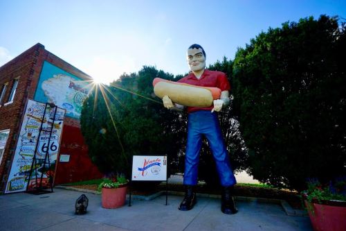A large statue of a man holding a hot dog stands outside a building, with trees and a bright sun in the background.