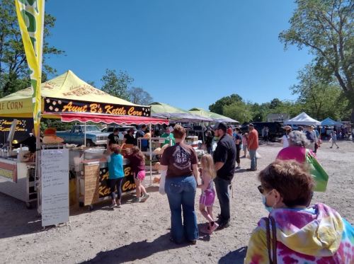 A busy outdoor market scene with people lined up at a kettle corn stand under sunny skies.