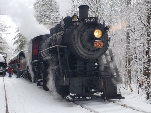 A vintage steam train in a snowy landscape, with smoke billowing and a person near the train.
