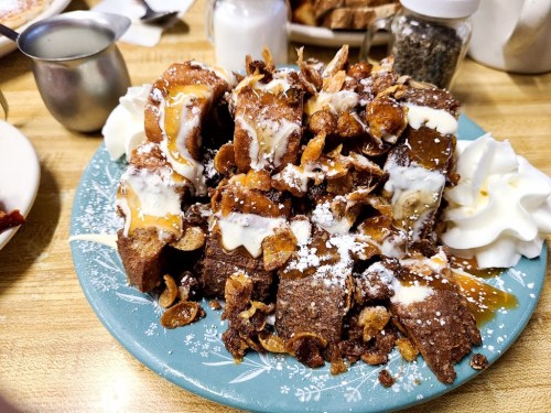 A plate of decadent dessert featuring chocolate bread, whipped cream, caramel drizzle, and crunchy toppings.