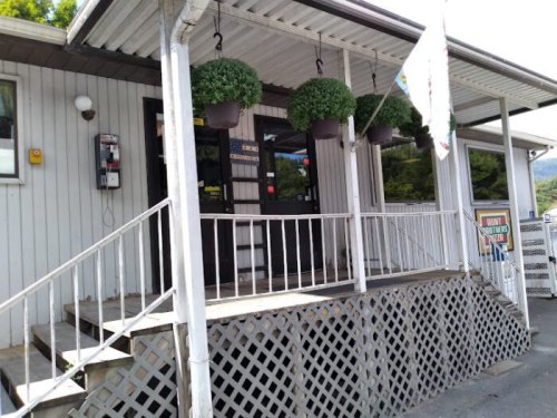 Entrance of a building with a wooden deck, potted plants hanging, and a phone booth nearby. Bright and inviting atmosphere.