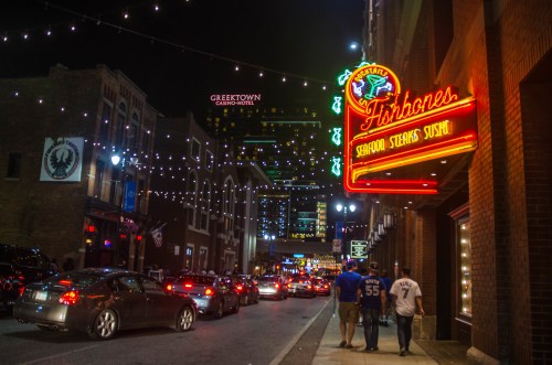 A bustling city street at night, lined with cars and illuminated by string lights and neon signs.