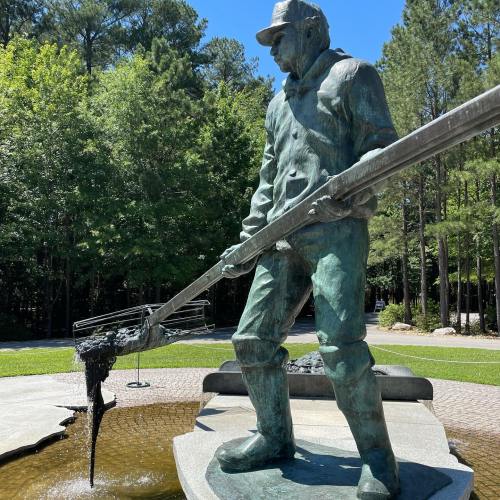 A bronze statue of a man in work attire, holding a long tool, surrounded by trees and a landscaped area.