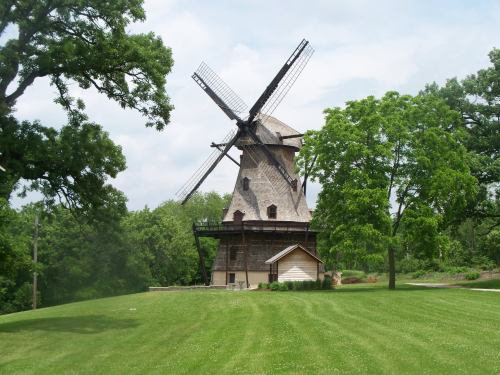 A historic windmill stands on a grassy hill, surrounded by trees under a cloudy sky.