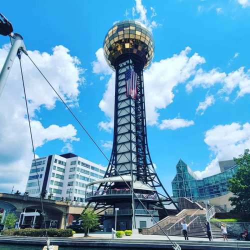 A tall observation tower with a golden dome, surrounded by blue skies and clouds, near modern buildings.